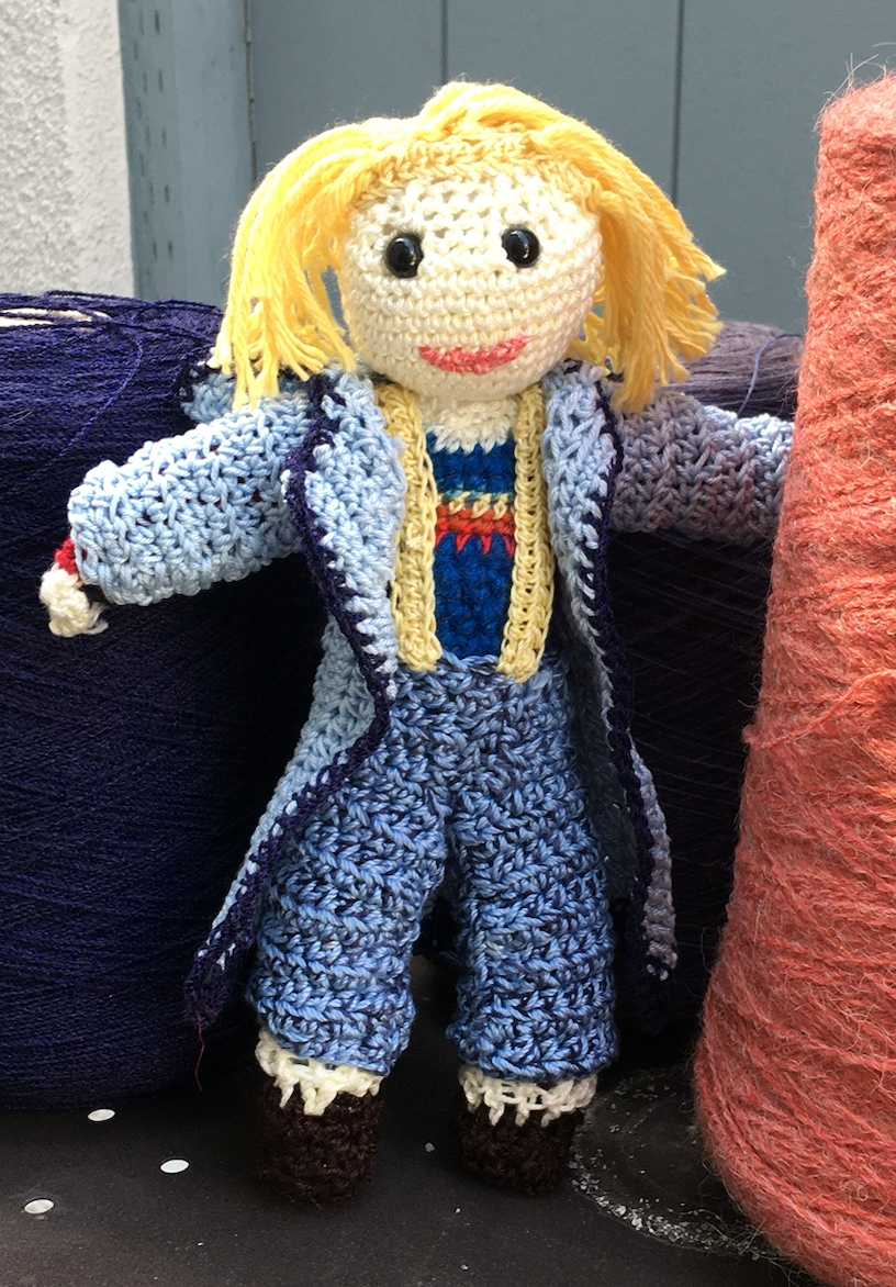 A crocheted doll resembling the Thirteenth Doctor from Doctor Who, with yellow hair, a blue coat, a rainbow t-shirt, yellow suspenders, and blue trousers